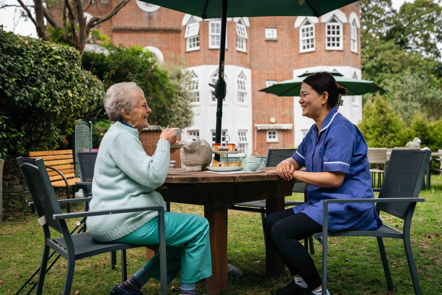 A resident and a carer sharing afternoon tea at a garden table, the white bay wing of the house behind them