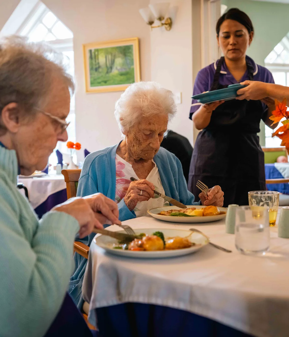 Two residents eating lunch at a table laid with white cloths while a carer serves