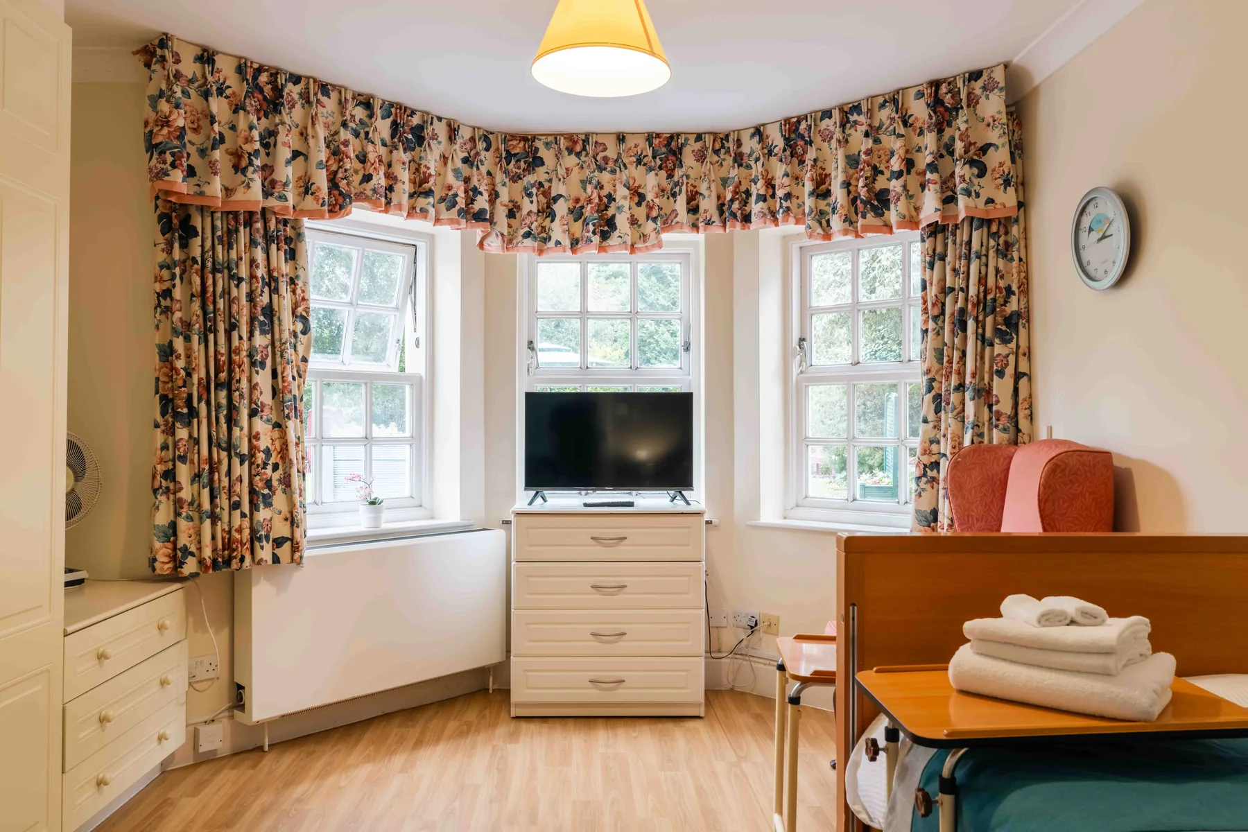 A bay-window bedroom with floral curtains, fresh towels set out on the bed table