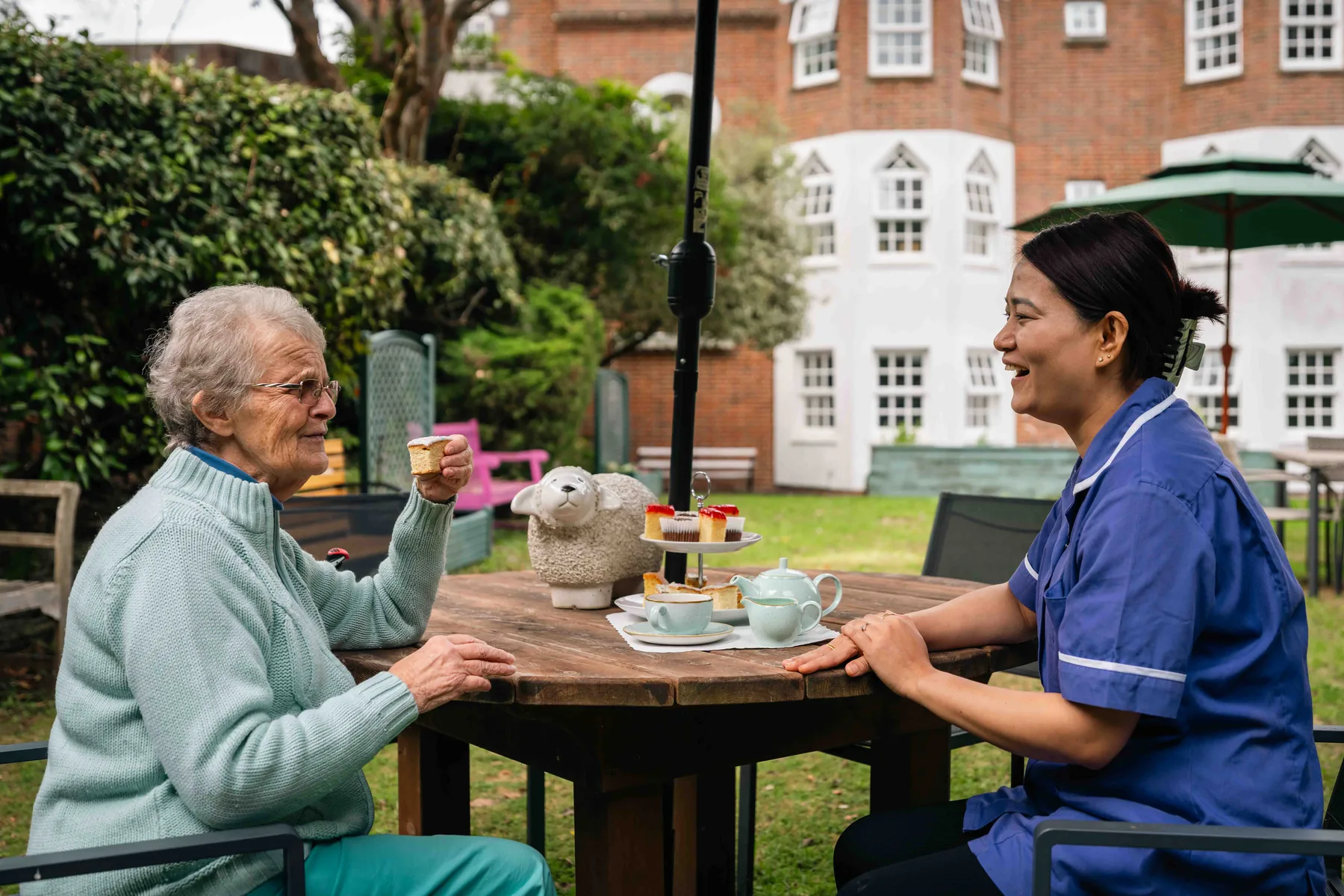 A resident and a carer laughing together over afternoon tea at a garden table